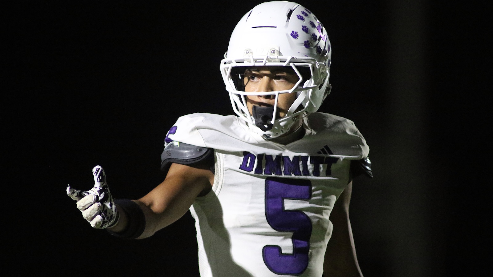 Football player in a white uniform with purple number 5, pointing with his left hand during a game, helmet decorated with purple paw prints.