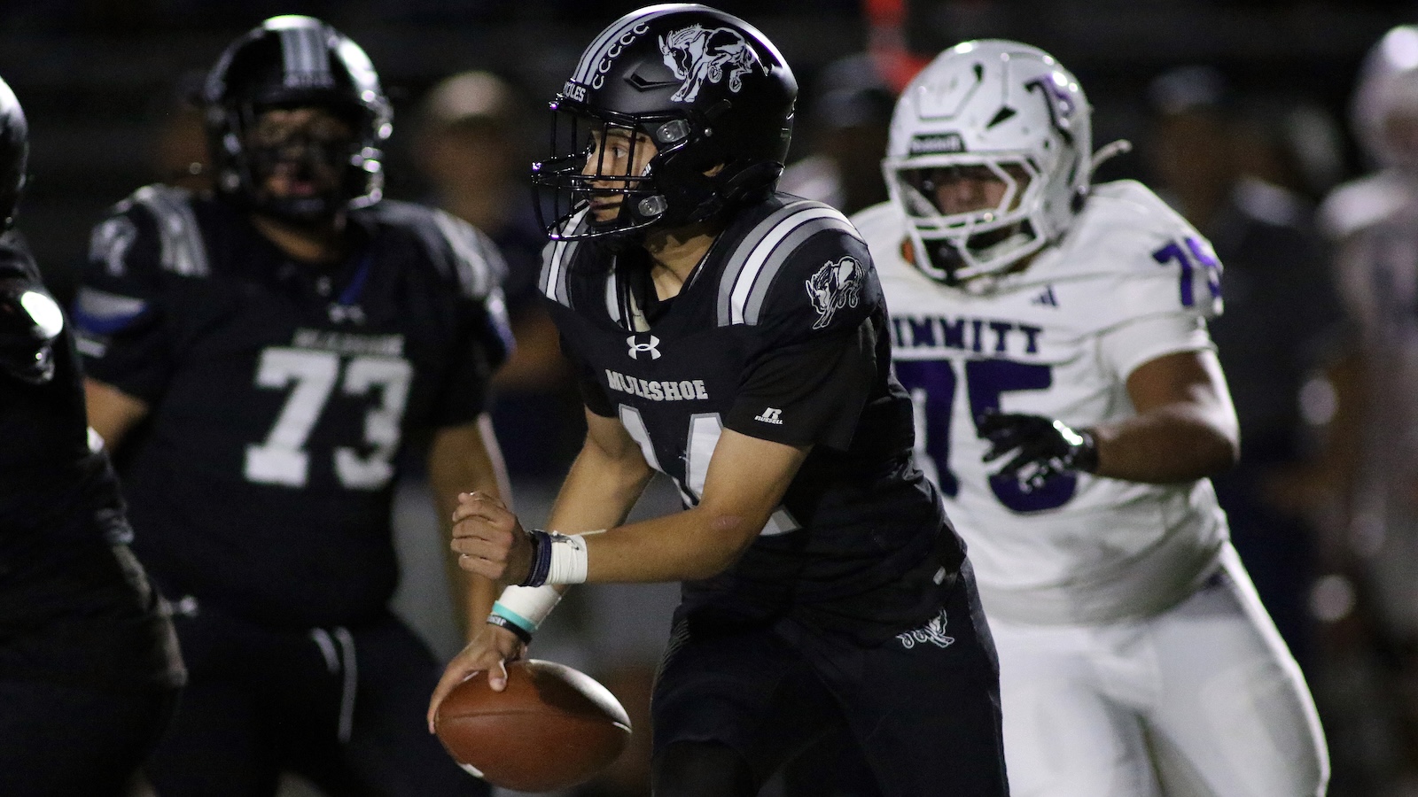 Football player in a black MULSHOE jersey holding the ball and running while defenders in white pursue him during a night game.