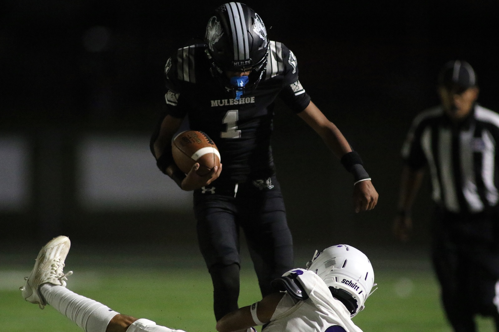 Running back in black #1 maintains ball while dodging a tackle from a white-uniform defender during a night football game under lights, a referee in the rear.
