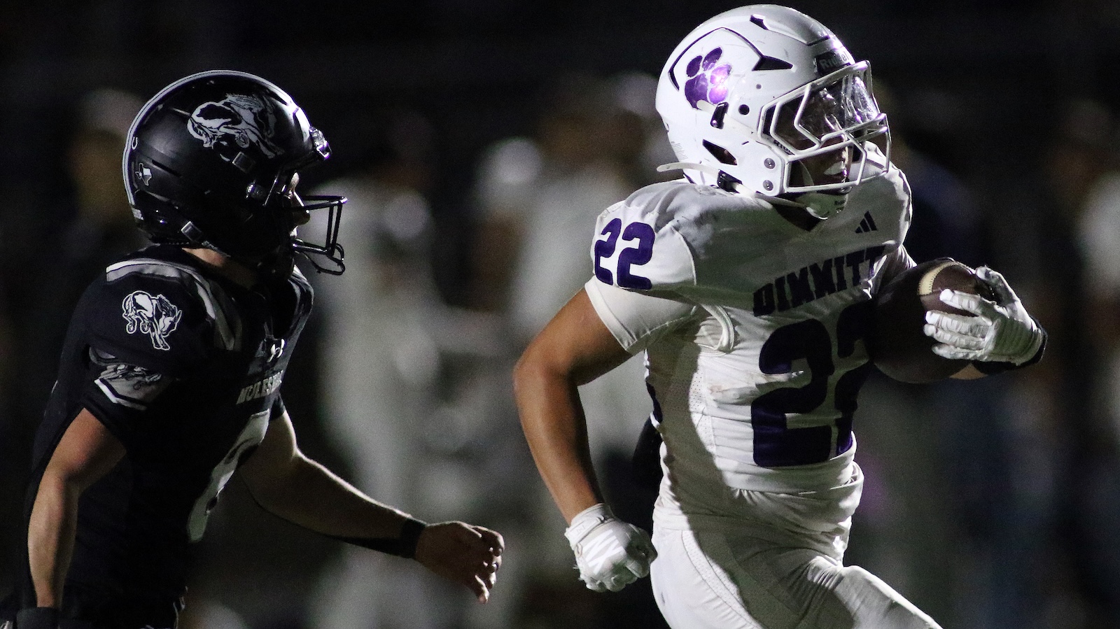 Football player in white jersey #22 runs with the ball, pursued by a defender in a black uniform at night.