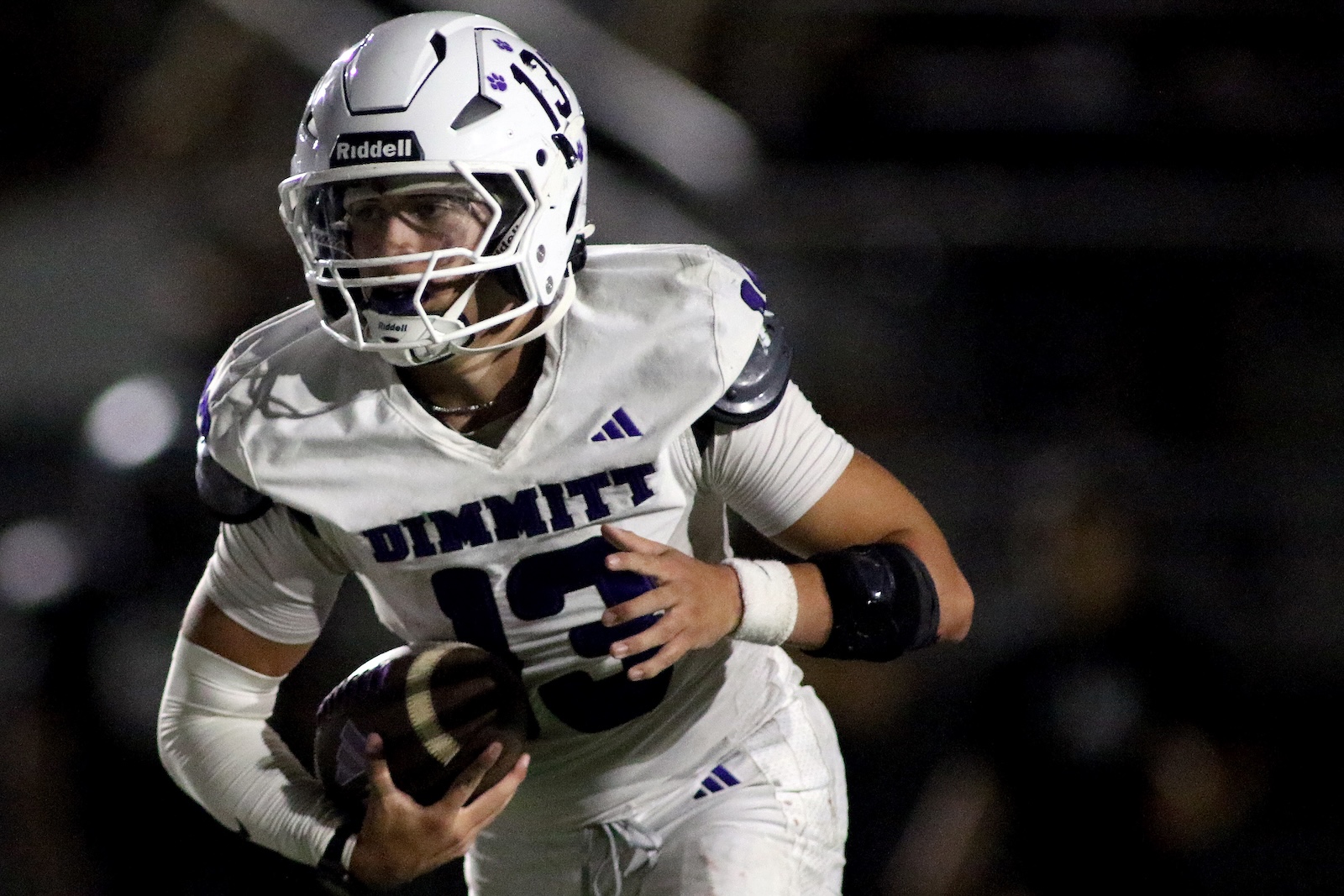 Football player in a white uniform and helmet, clutching the ball and running on a dark field during a night game.