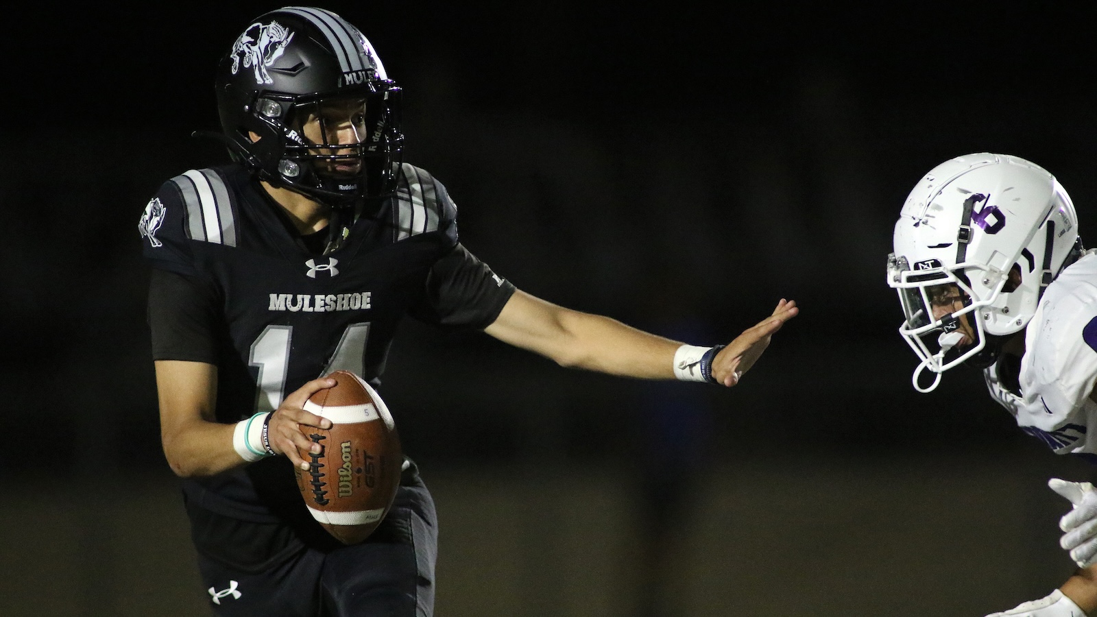Quarterback in a black MULESHOE jersey holds a football and reaches out to stiff-arm a white-helmet defender during a night game.