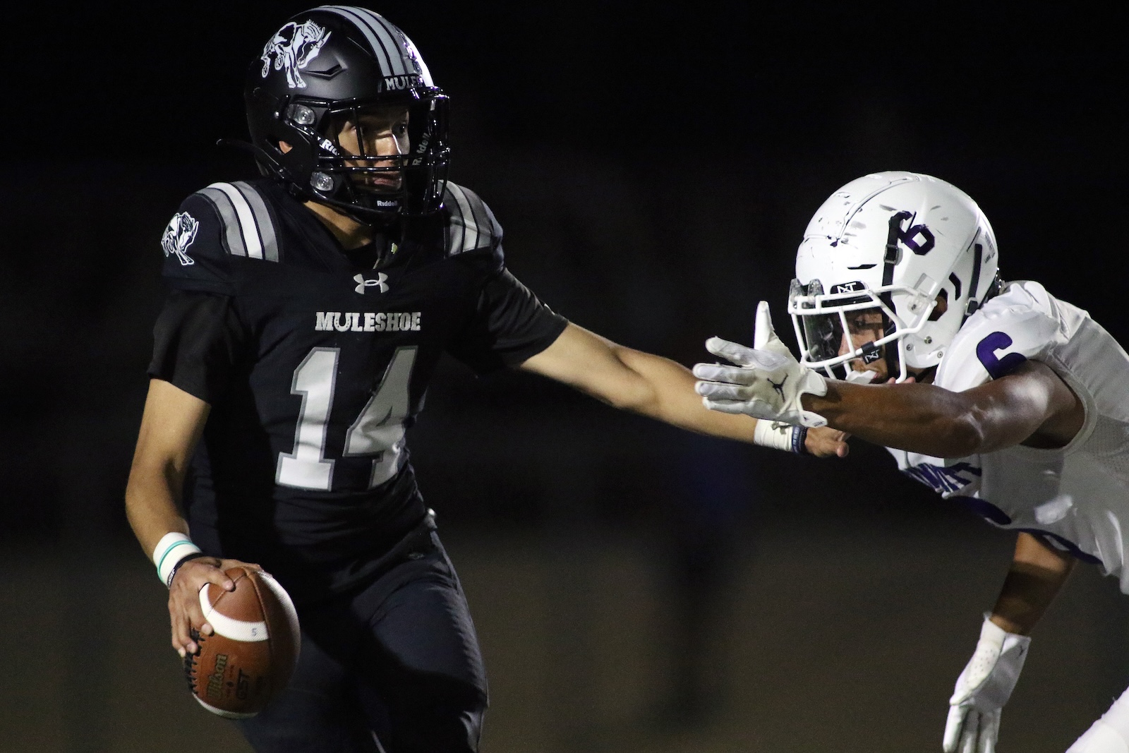 Muleshoe football player in black with #14 holds the ball and reaches past a white-uniform defender at night.