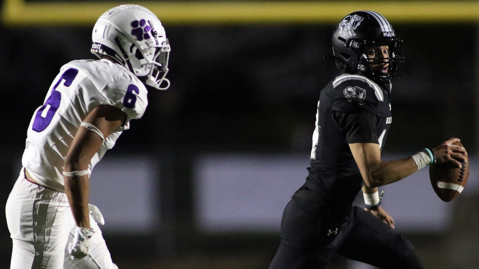 Quarterback in black runs with the football while a white-uniform defender with purple 6 pursues him at night.