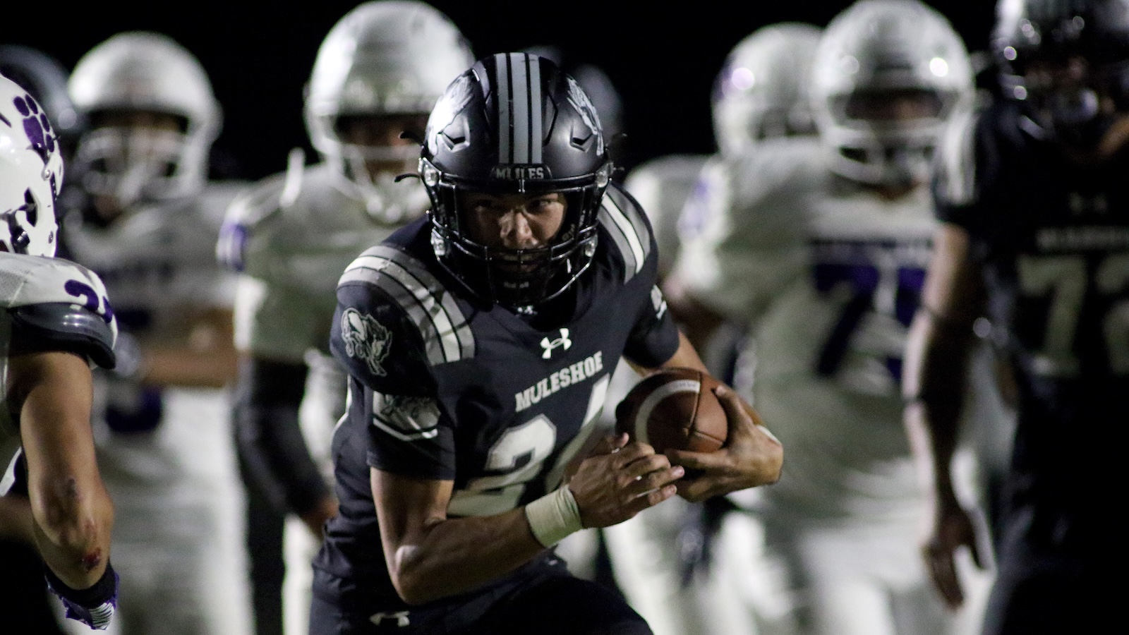 Male football player in a dark uniform runs with the football during a night game, surrounded by defenders in white uniforms.