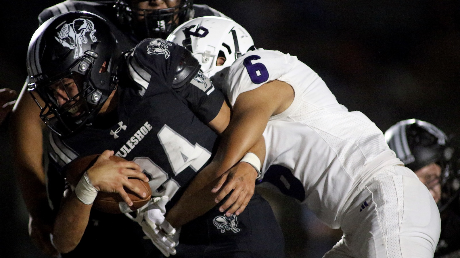 Football action: a ball carrier in a dark jersey is tackled by a white-uniform defender at night, gripping the ball tightly.