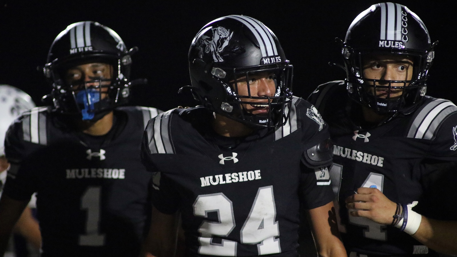 Three Muleshoe High School football players in black uniforms and striped helmets stand together on a night game field, centered on #24 player smiling veering forward out of the line-up.