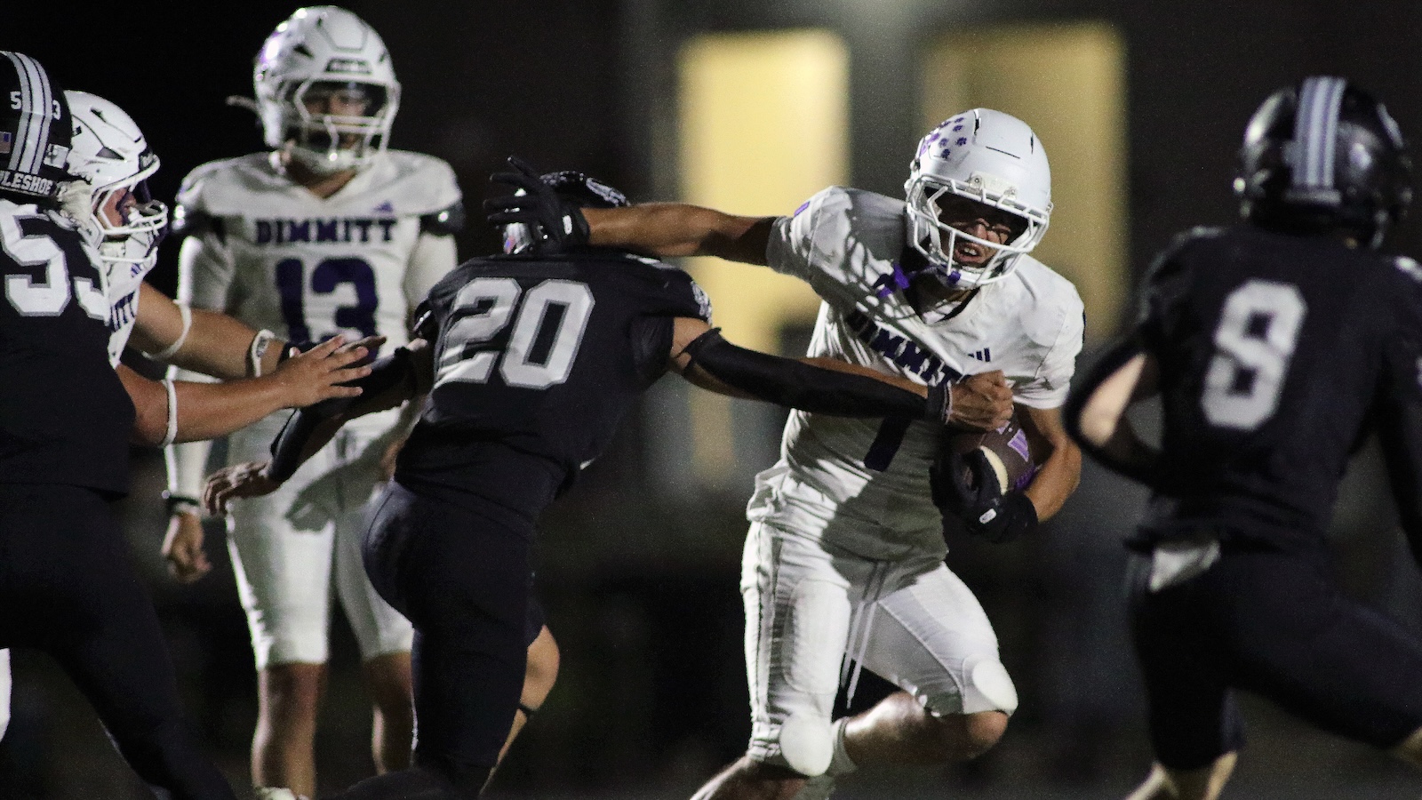 Night football game: a white-uniform running back is tackled by a black-uniform defender while teammates look on.