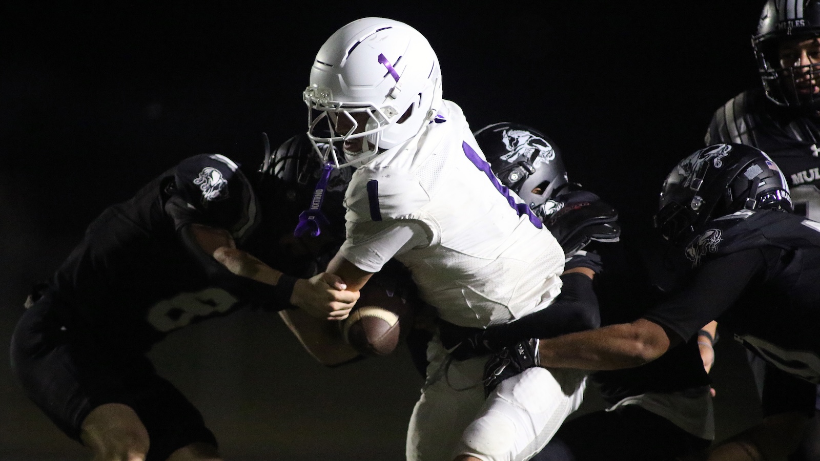 Night football action: a white-uniformed player with purple trim runs with the ball as defenders in dark uniforms grab at him under stadium lights in the background.