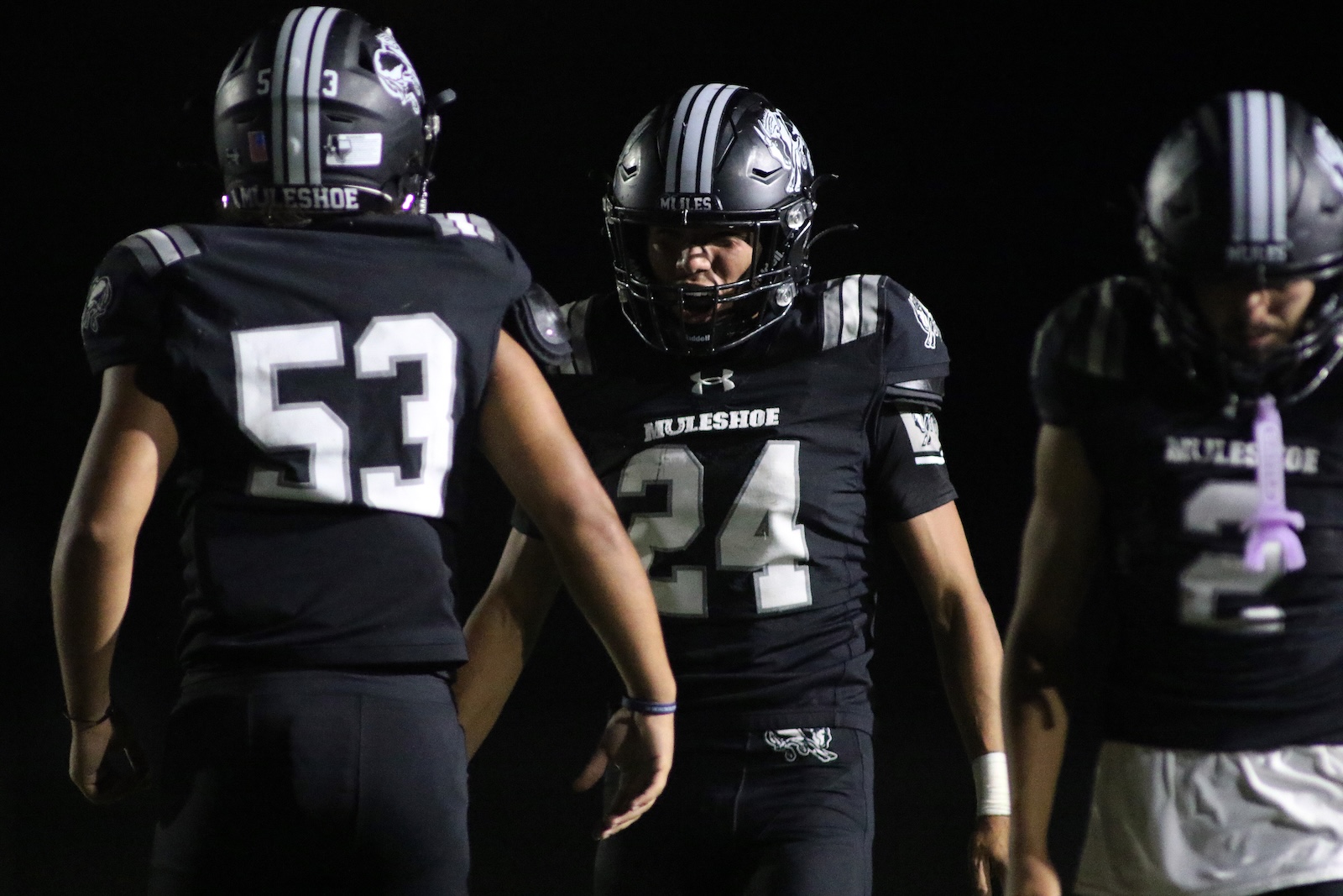 Three football players in black uniforms with white numbers stand in a huddle at night, facing each other on the field (Muleshoe team).