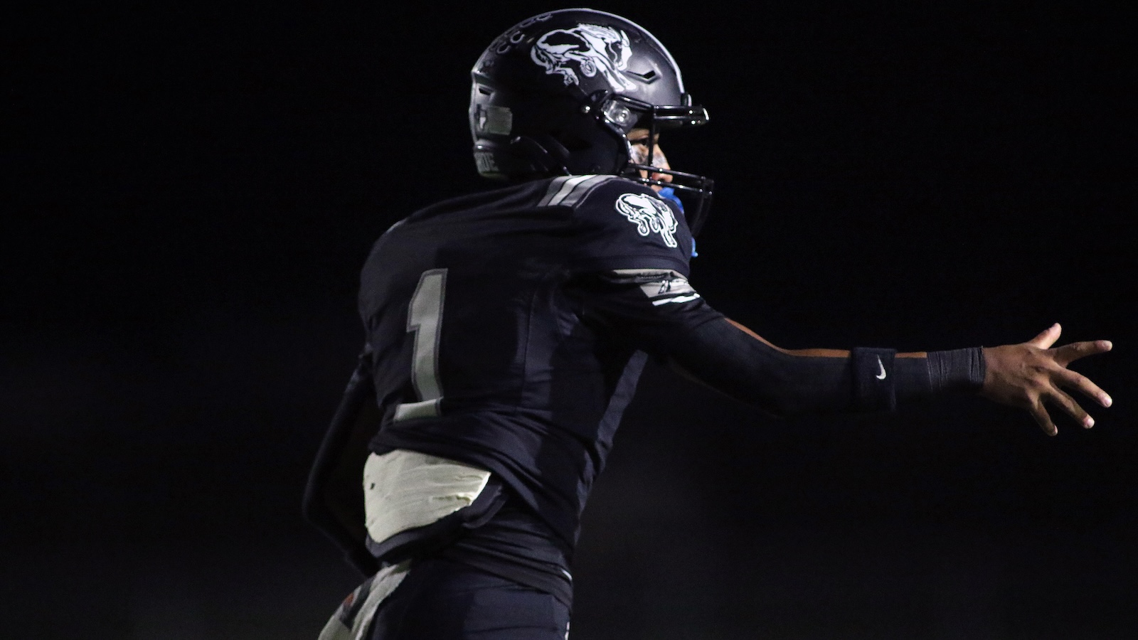 American football player in a dark uniform reaches forward during a night game, helmet logo visible