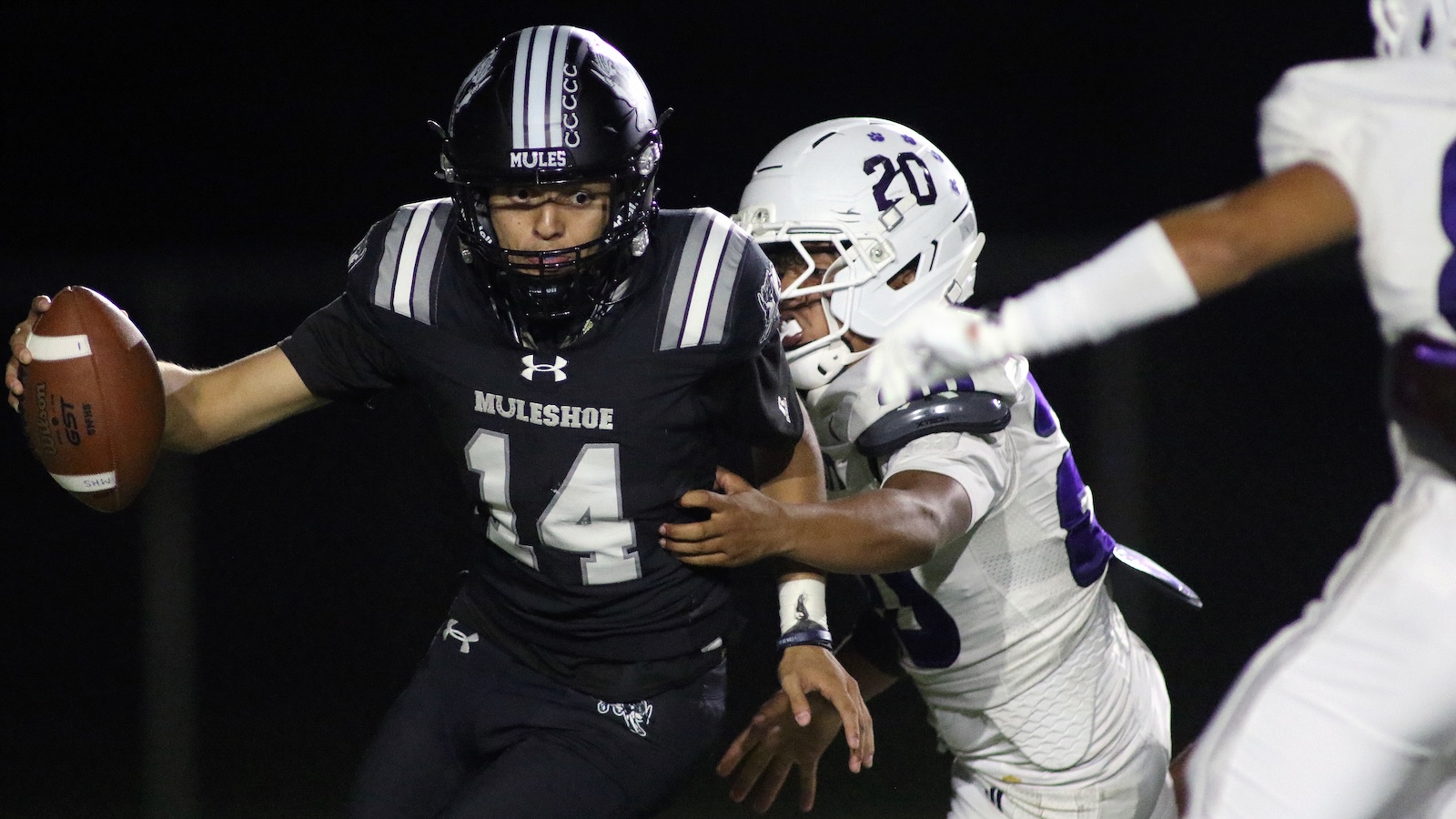 Muleshoe football player wearing #14 in black jersey runs with the ball at night, pursued by a white-uniform defender.