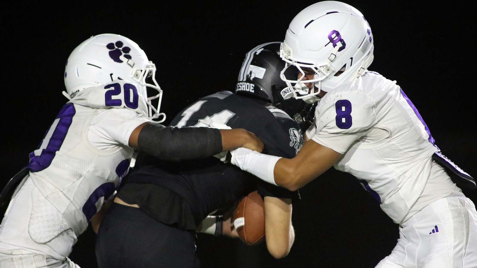Three football players collide: two white-uniformed players (numbers 20 and 8) tackle a dark-uniform ball carrier at night, holding the ball.