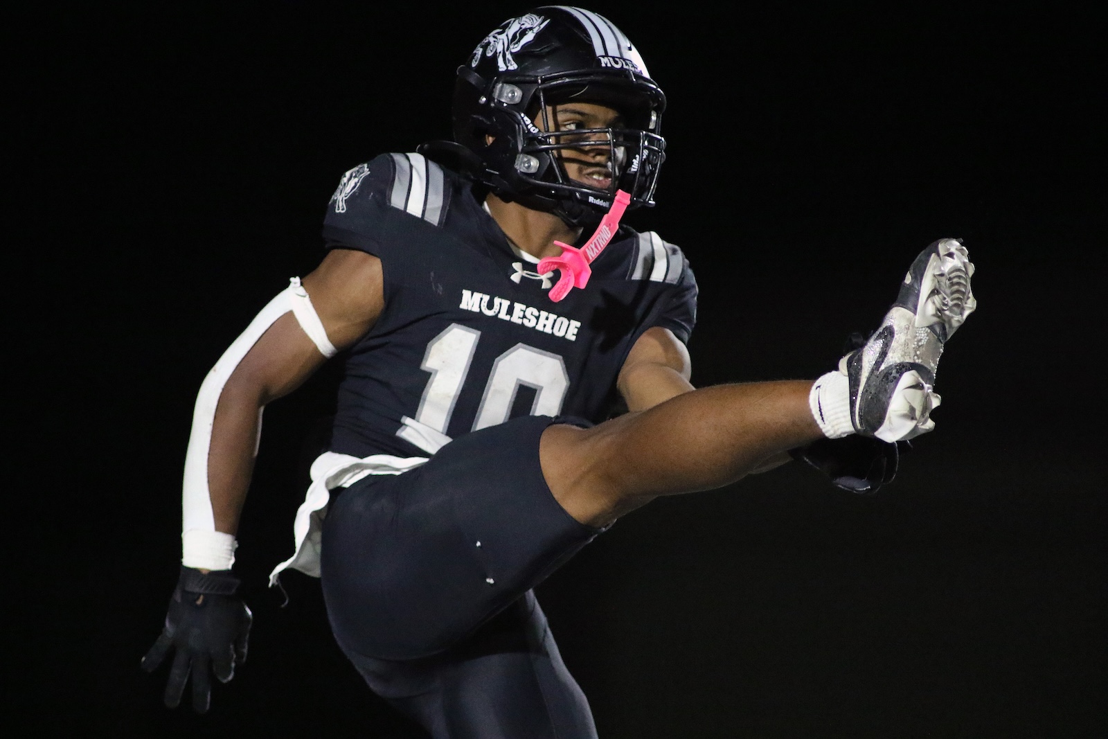 Football player in a black Muleshoe jersey (#10) delivering a high kick mid-air during a night game.