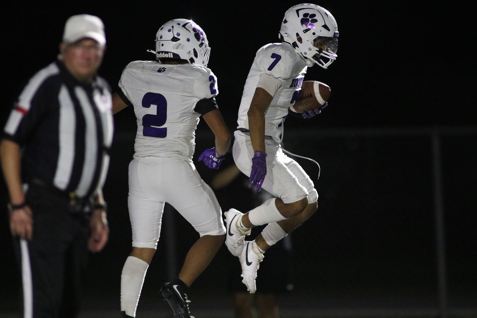Nighttime football play: a running back (number 7) carries the ball while a teammate pursues nearby; referee left in view.