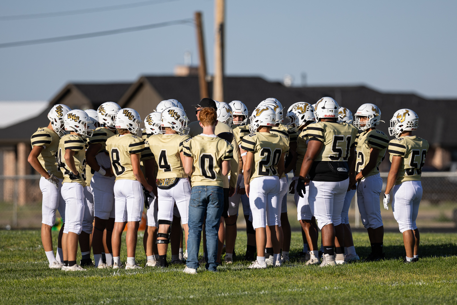 Youth football team in gold jerseys huddles on a sunny field, coaches guiding them. (15 players in white pants and helmets)