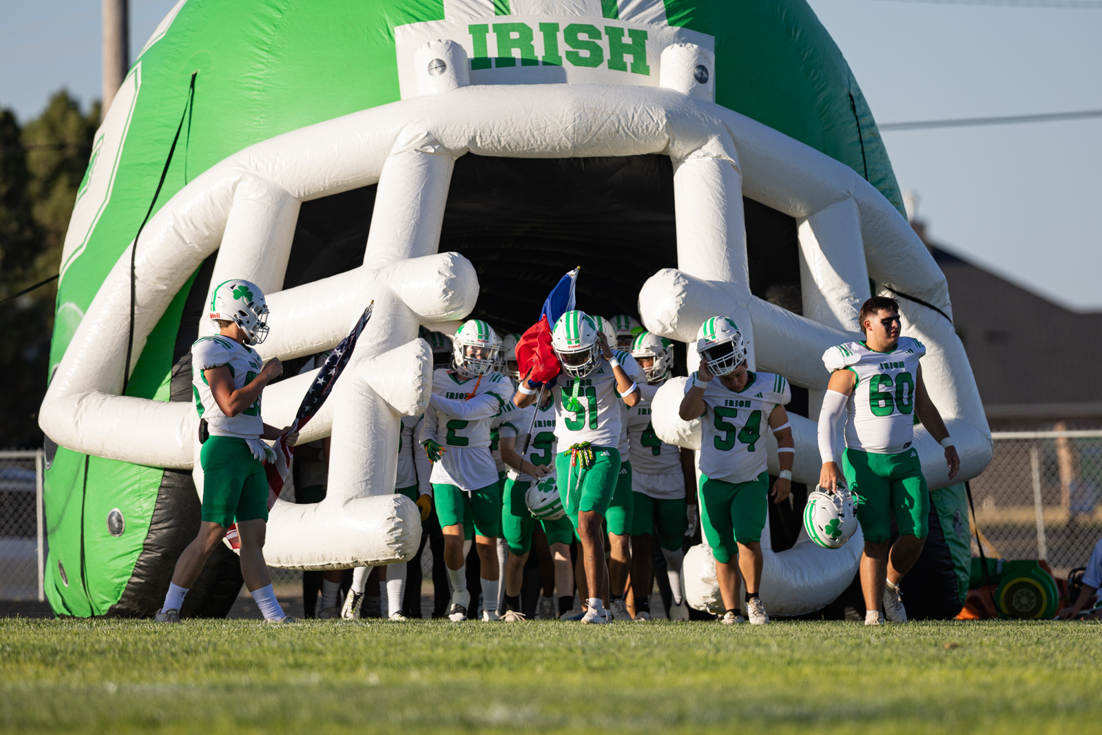 Youth football players in green and white uniforms rush through a large inflatable green tunnel labeled IRISH before a game.