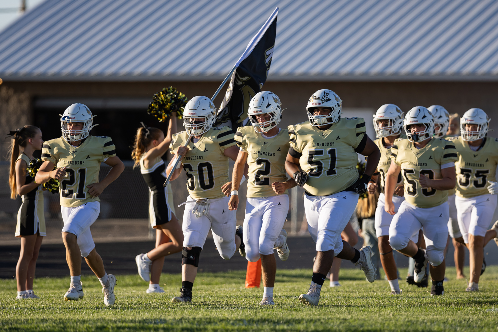 High school football team in gold jerseys runs onto the field, led by a flag, with cheerleaders following in the background.