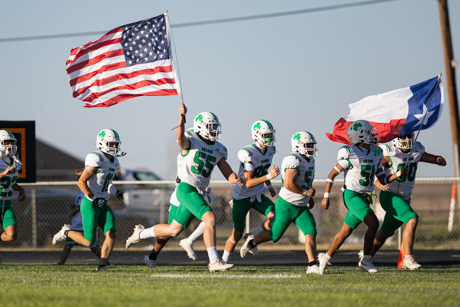 Football players in white-and-green uniforms run onto the field as one player raises the American flag and another carries a Texas flag.