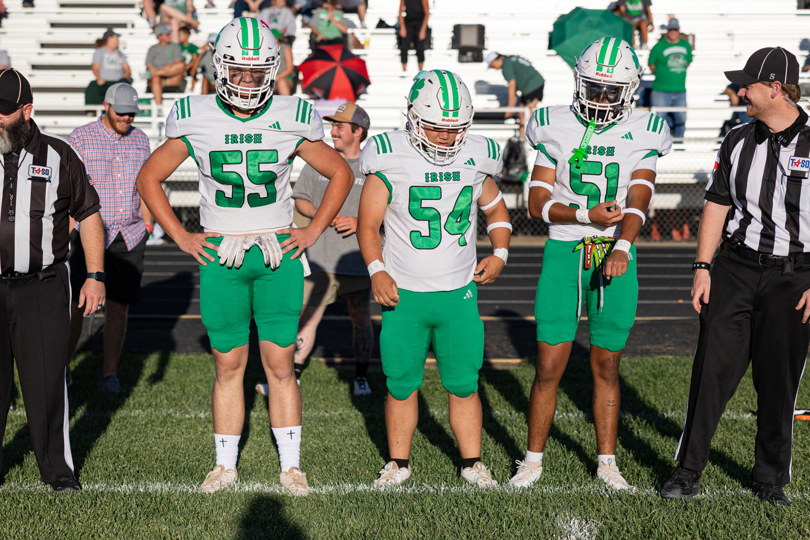Three Irish football players in white jerseys with green numbers 55, 54, and 50 stand with referees on a football field before a game.
