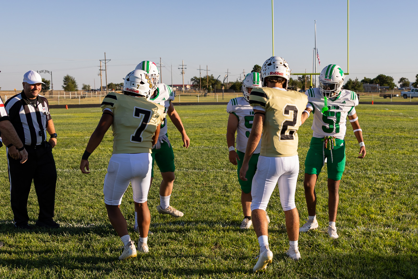 Youth football players on a grassy field during a game, two teams facing off near a goalpost with a referee watching nearby.