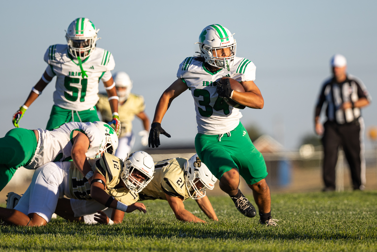 Football game: a player in white/green jersey with the ball runs forward while opponents dive on the grass in front, referee in background.