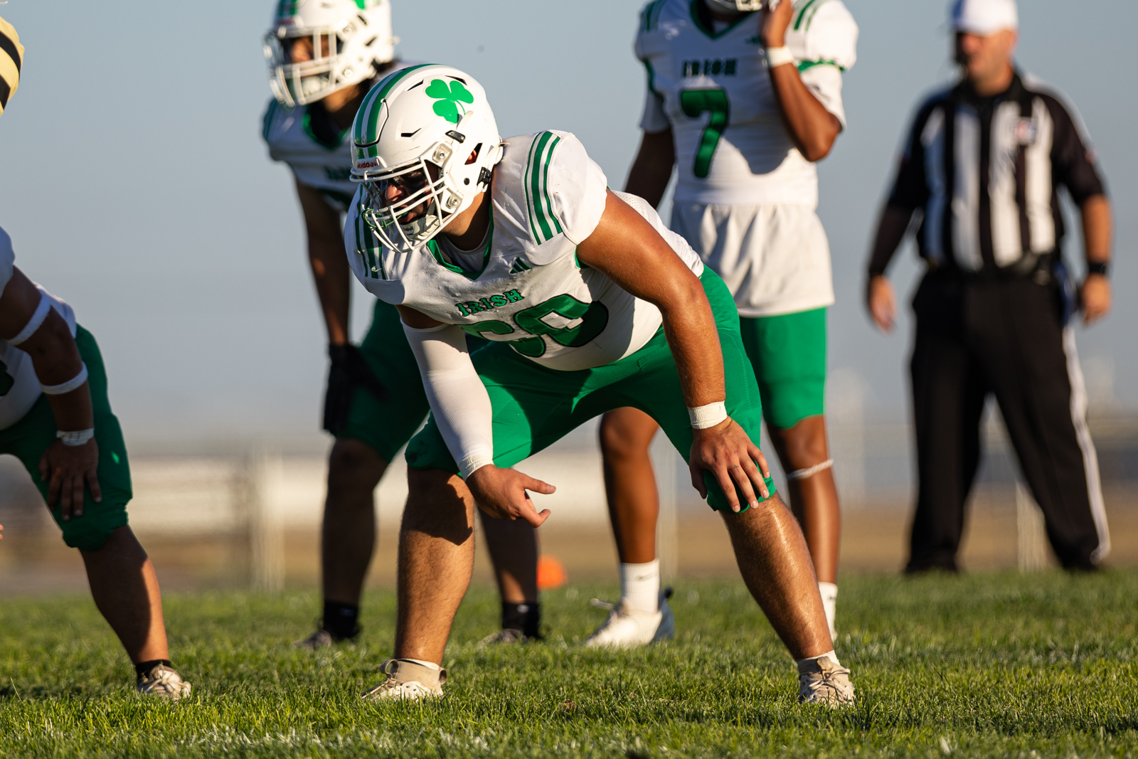 Football lineman in a green-and-white uniform crouches at the line of scrimmage, ready for the snap on a grassy field, with teammates nearby.