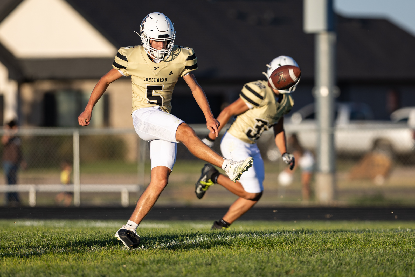 Football players in gold uniforms on a sunny field; #5 is kicking the ball as a teammate follows nearby.