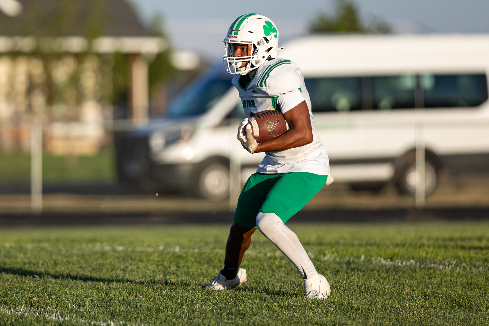 Football player in white jersey and green shorts running with the ball on a grassy field during a game.