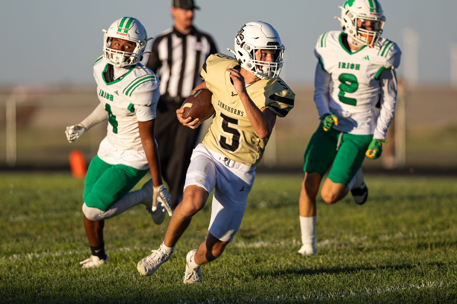 Football player in a gold Longhorns jersey (number 5) runs with the ball while two defenders in white and green chase him on a grassy field.