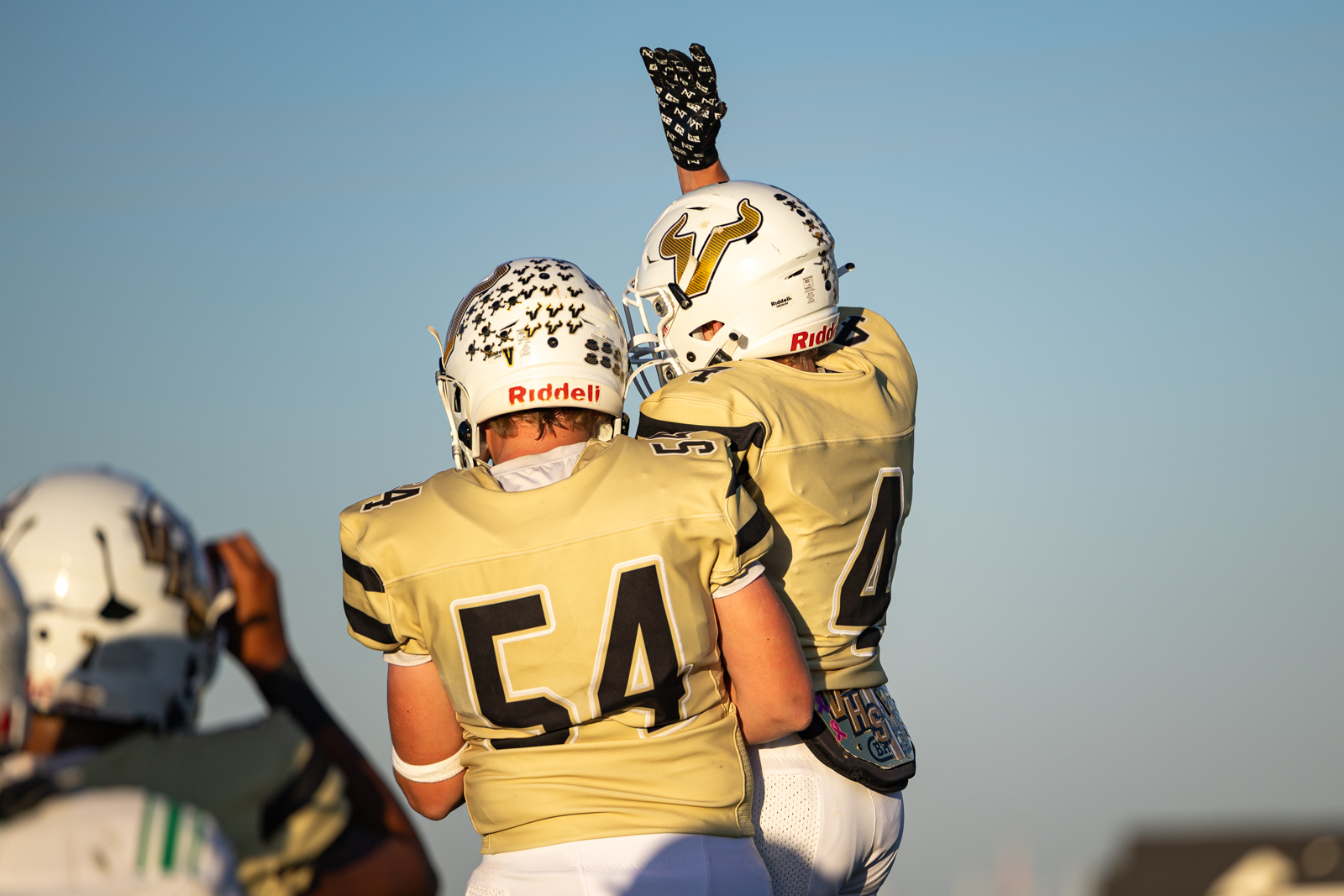 Two football players in gold jerseys embrace as one raises a gloved hand in celebration against a clear sky backdrop.