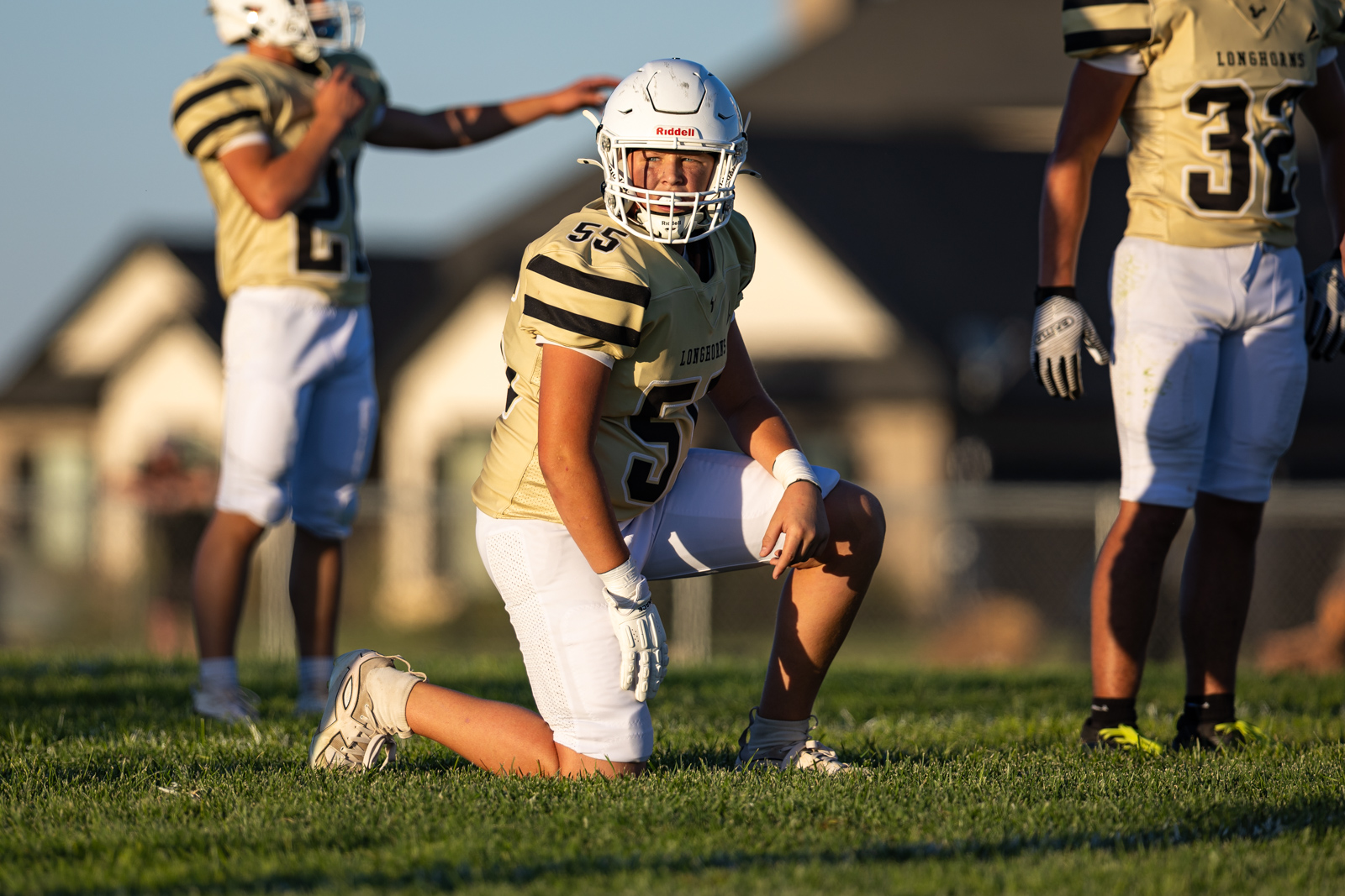 Football players in gold jerseys kneel on a sunny field; central player wears white helmet with number 55 and'Longhorns' on the jersey.