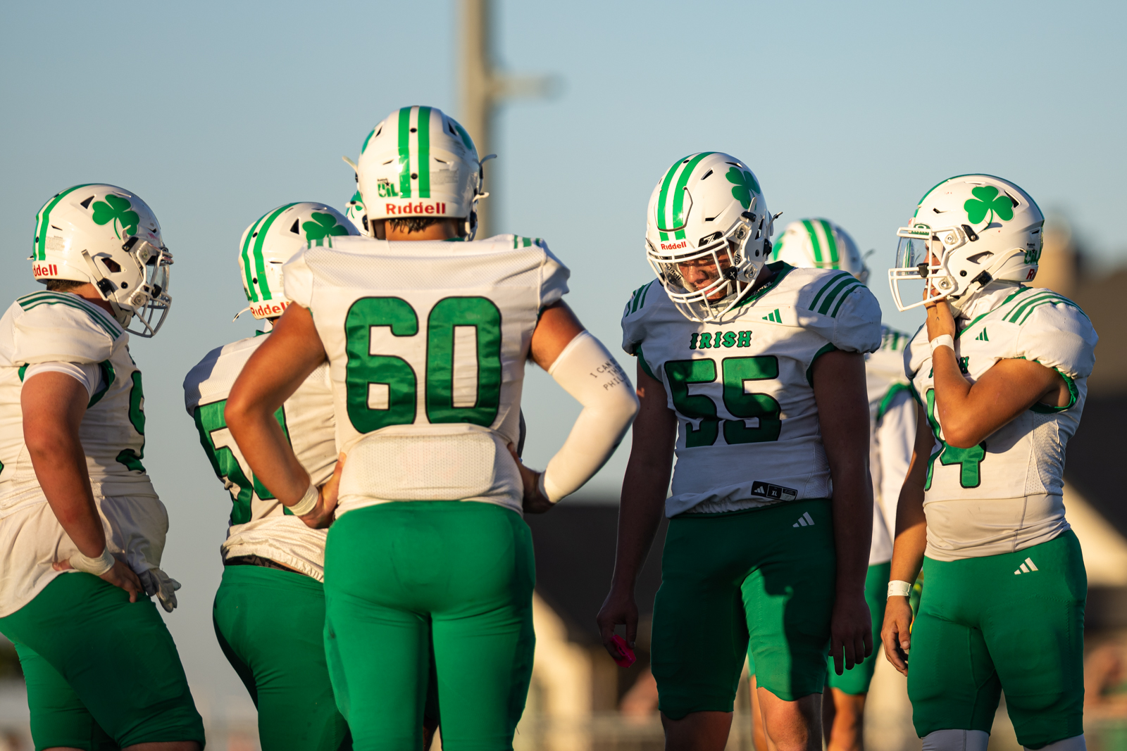 Group of Irish football players in white and green uniforms huddle on the field during sunset, helmets with shamrocks visible.