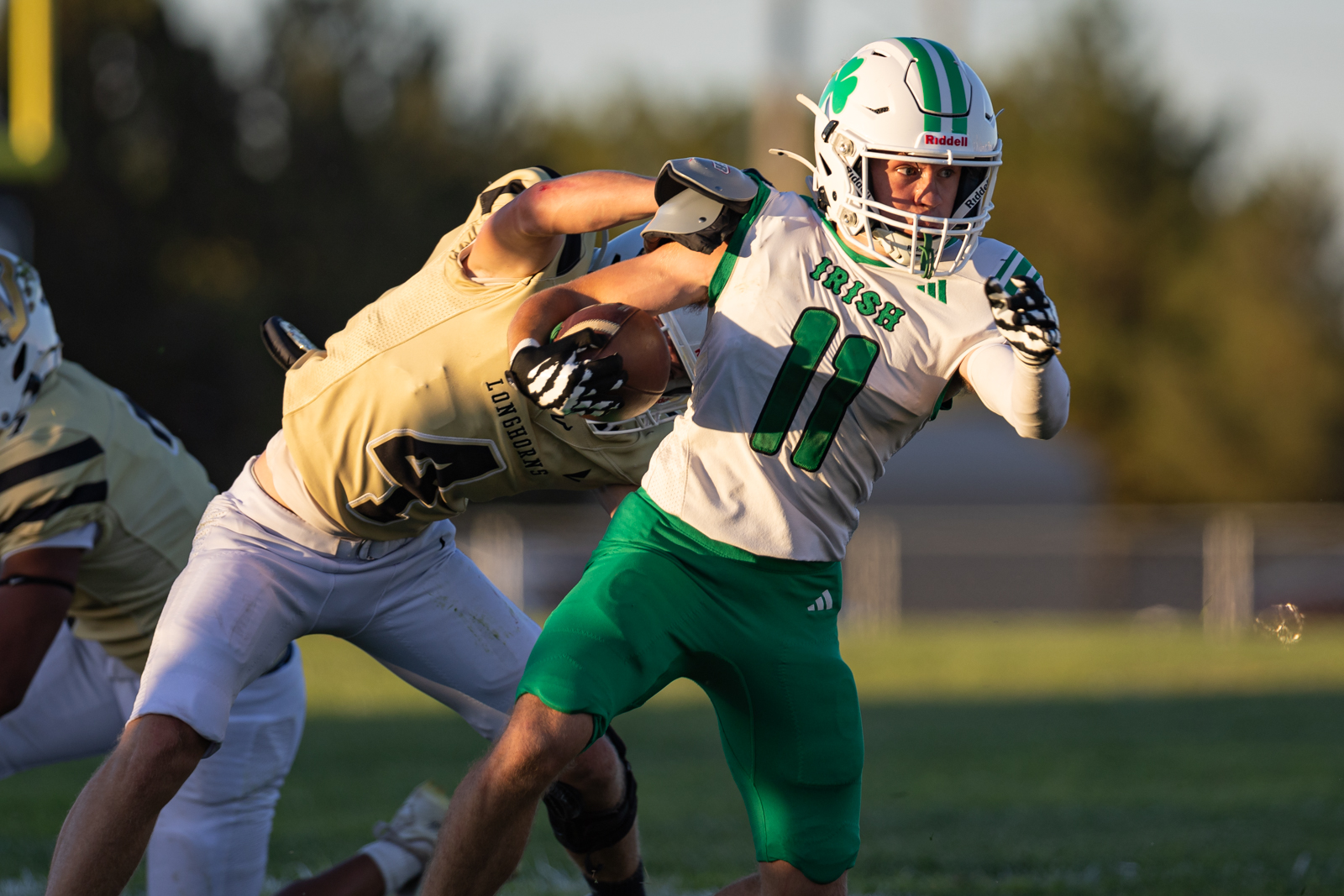 White-and-green football player (#11) runs with the ball as a defender in tan jersey dives in for a tackle on a sunny field.