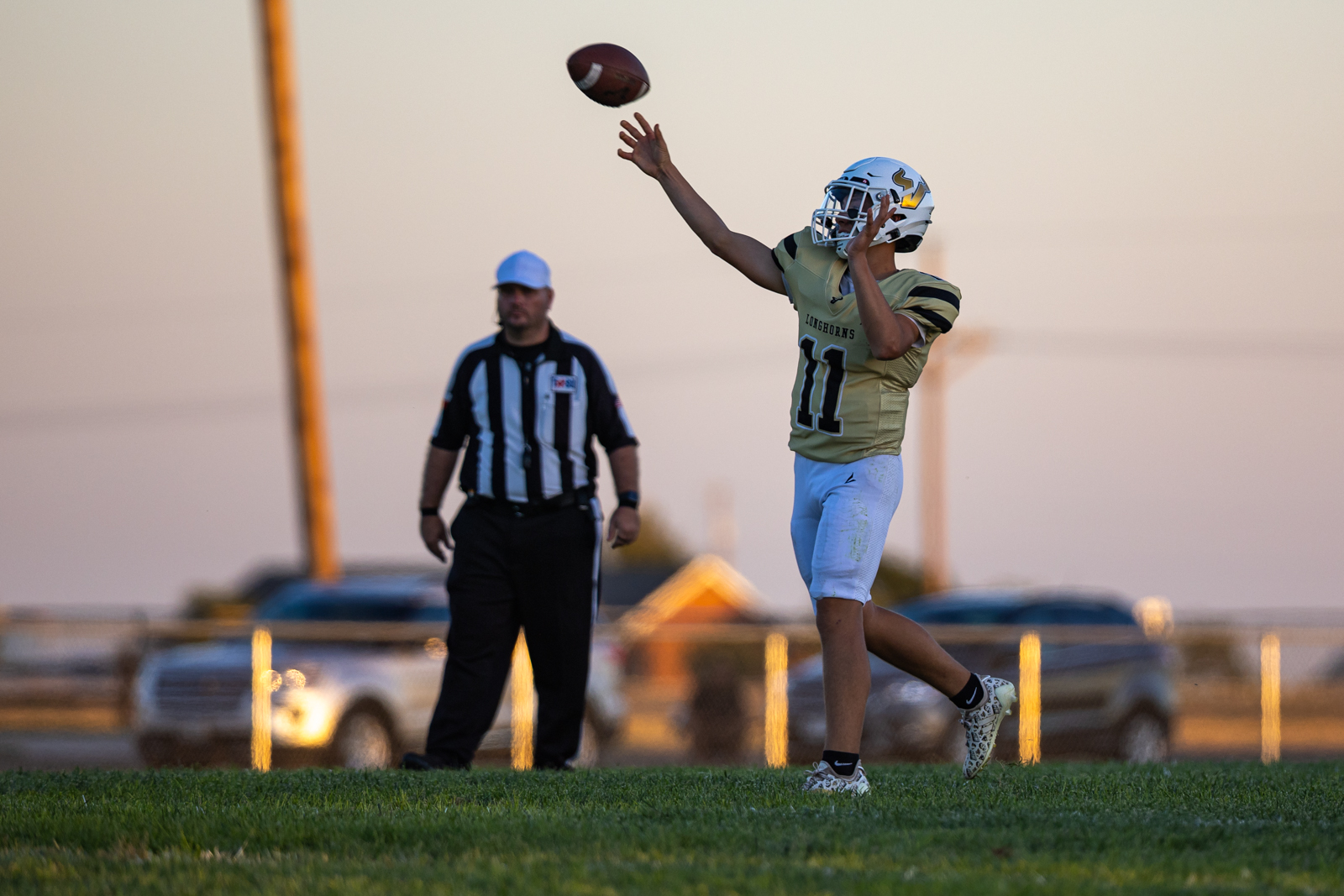 Football player in gold No. 11 leaps to catch a pass, as a referee watches in the background at sunset distance.