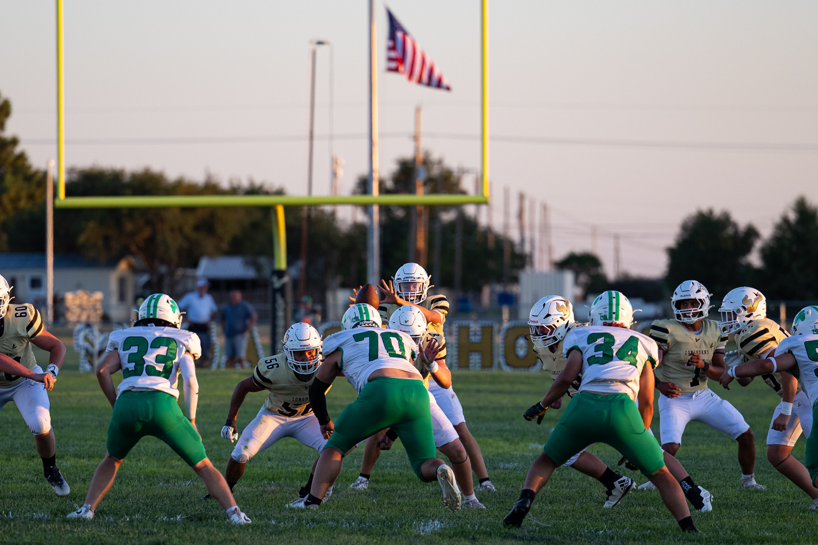 Quarterback throws a pass while teammates block at the line of scrimmage on a sunny football field.