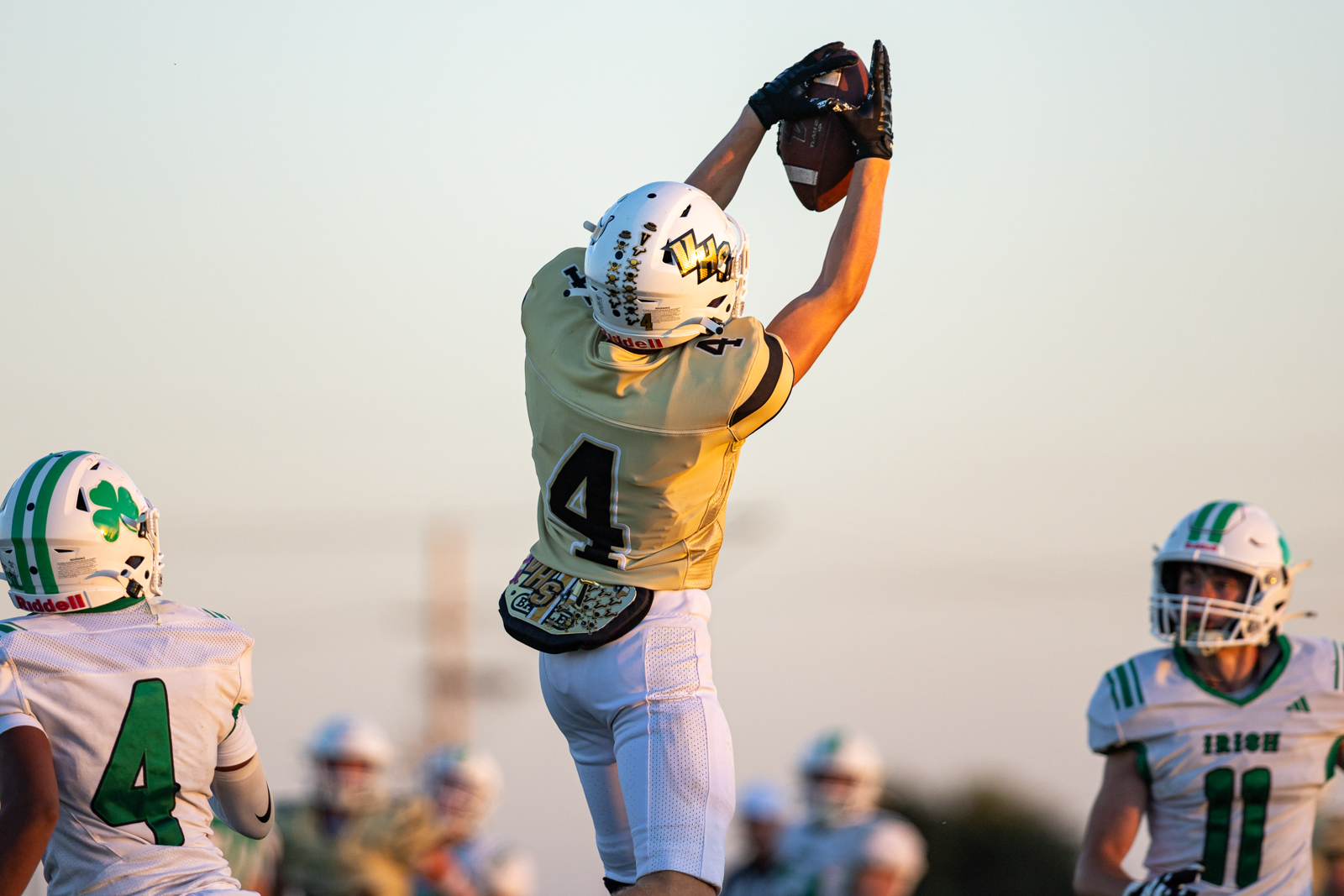 Football player in a tan/gold jersey (#4) jumps to catch a pass, gloves raised, with teammates in white and green nearby.