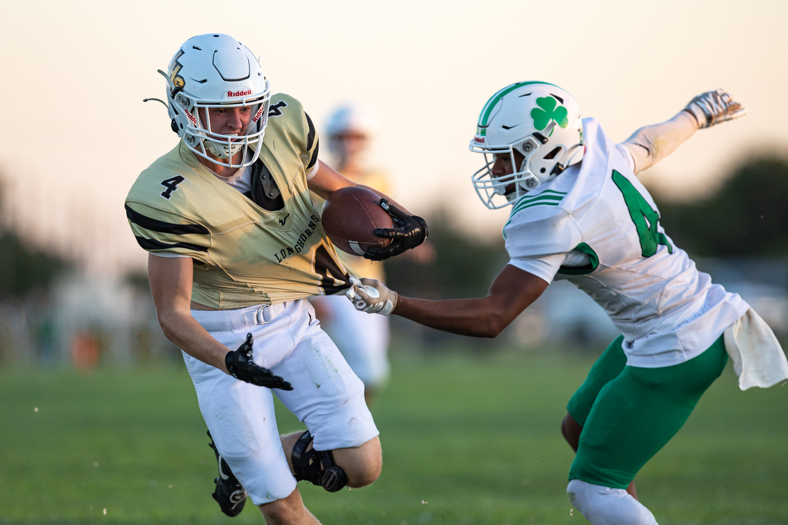 Football players in a tense moment: ball carrier in tan jersey dodges a tackle on a grassy field at sunset.