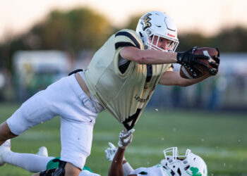 SHS-VHS-FB-28 | Press Pass Sports Football player in a gold jersey and white helmet lunges forward to catch a ball, reaching over a sprawled opponent on a grassy field.
