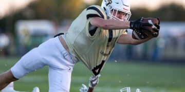 Football player in a gold jersey and white helmet lunges forward to catch a ball, reaching over a sprawled opponent on a grassy field.
