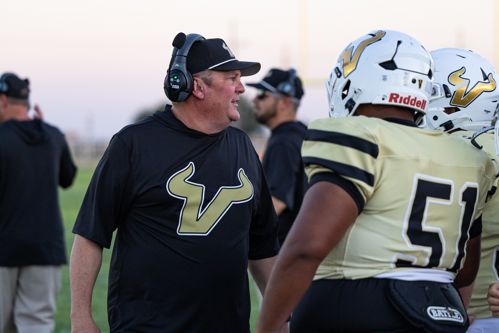 Coach wearing a headset talks to football players on the sideline during practice on a sunny day