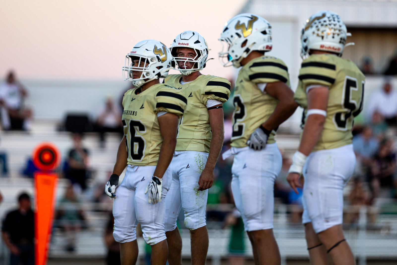 Football players in gold jerseys and white helmets stand on the sideline during a game, numbers 26 and 2 visible, orange yard marker in the background
