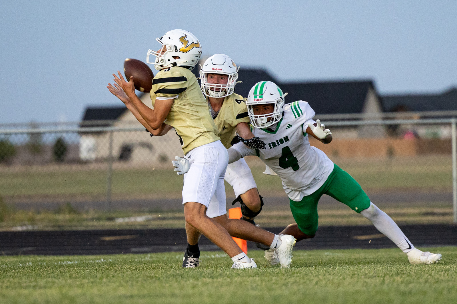 Quarterback in a gold jersey prepares to throw while two defenders in white/green close in on him on a football field.