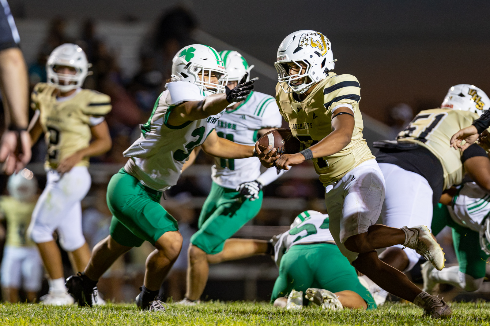 Football player in gold advances with the ball as green-clad defenders pursue on a grassy field at night under stadium lights.