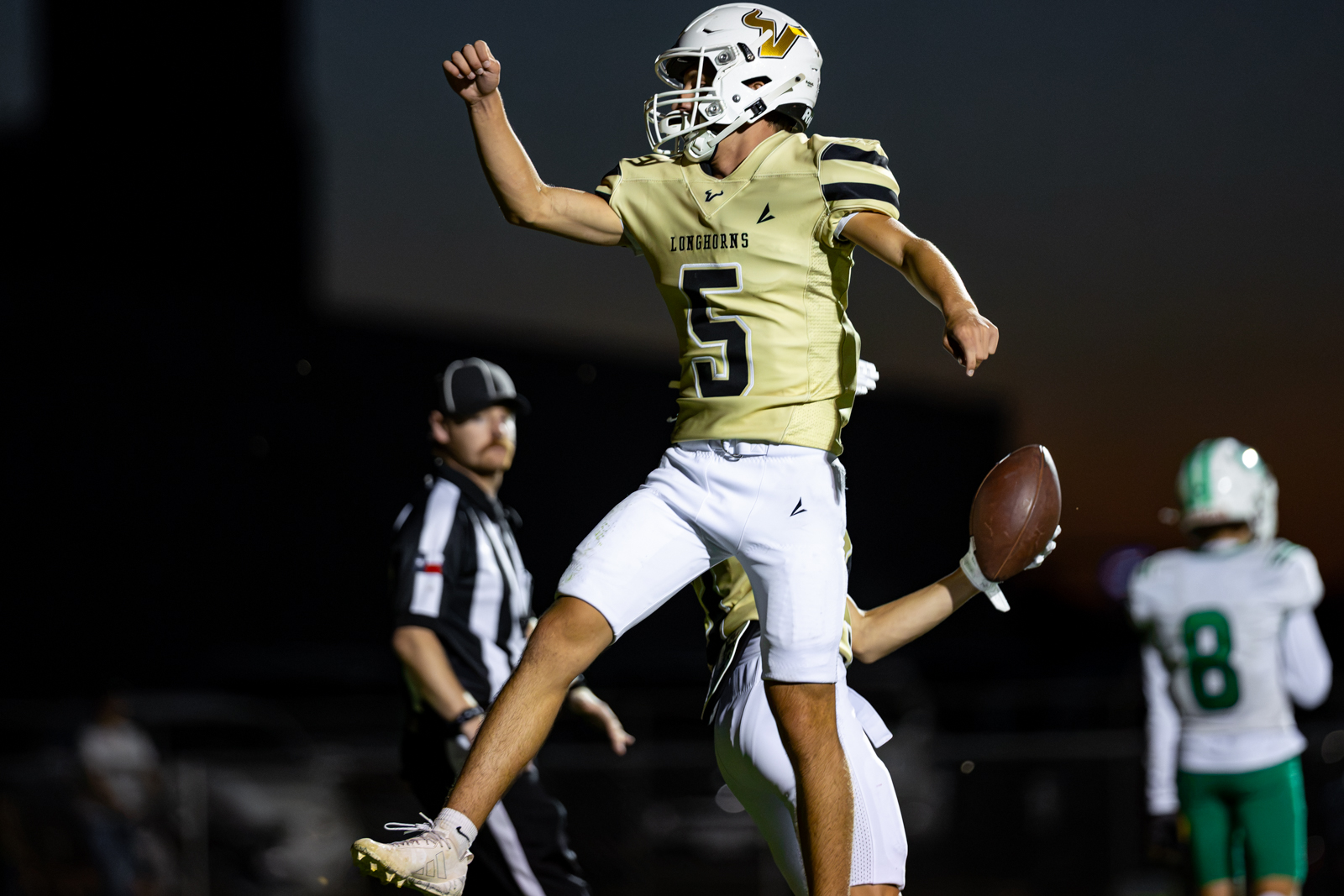 Football player in a gold Longhorns jersey (#5) leaps in the air with arms raised during a game, a referee and an opposing player in the background, nighttime stadium scene