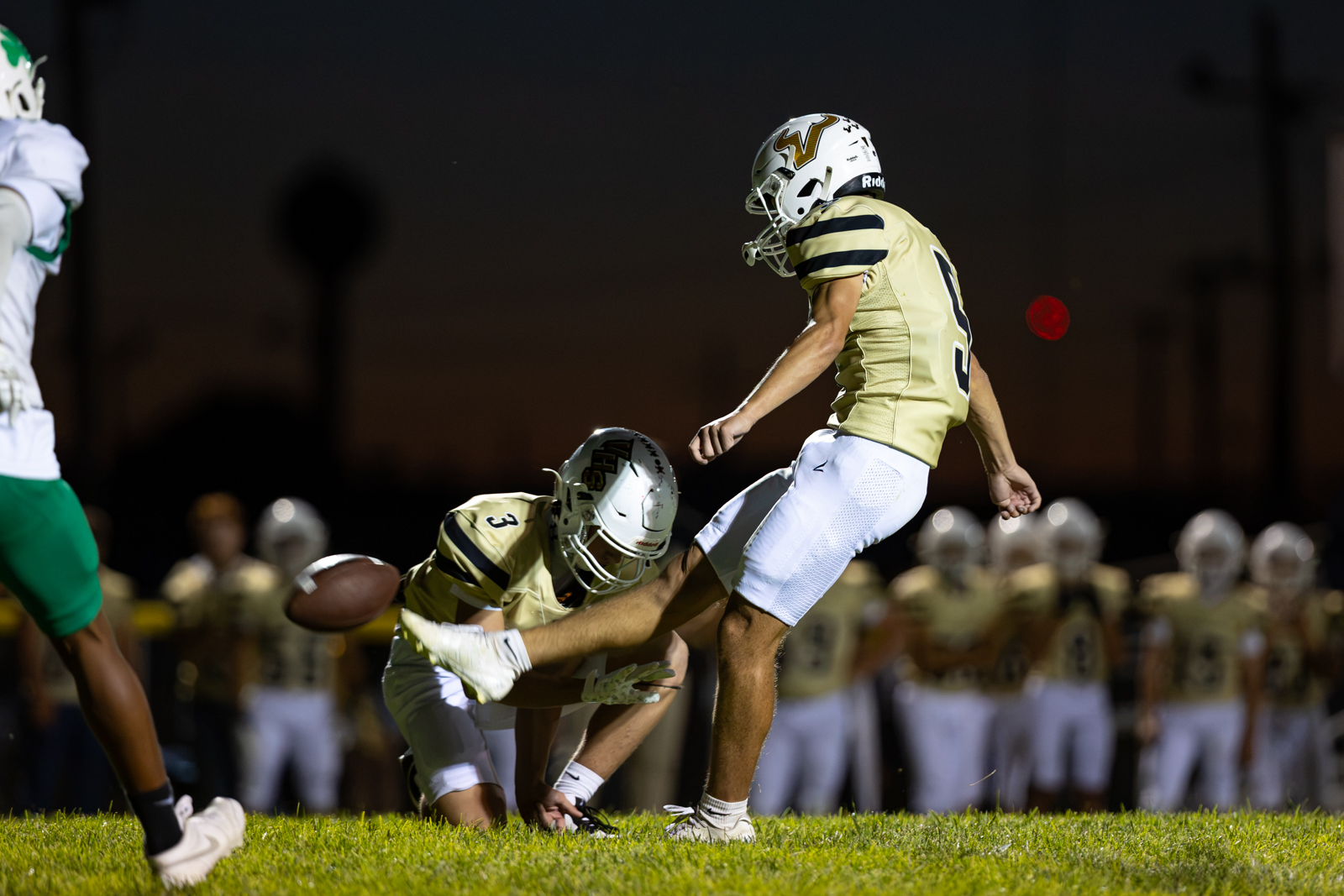 Nighttime football scene: players in gold uniforms pursue a loose ball on the field as a kicker and a teammate converge near the ball.