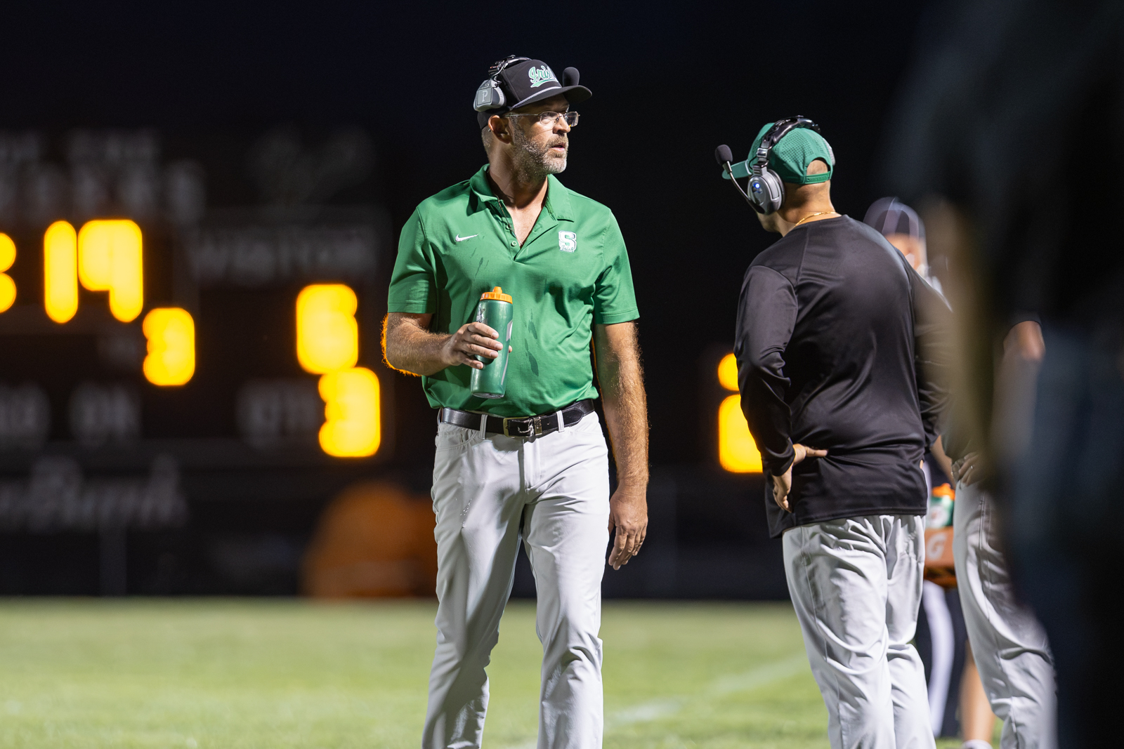 Football coach in a green polo and cap holding a water bottle, speaking to a teammate on a sideline at night under stadium lights.