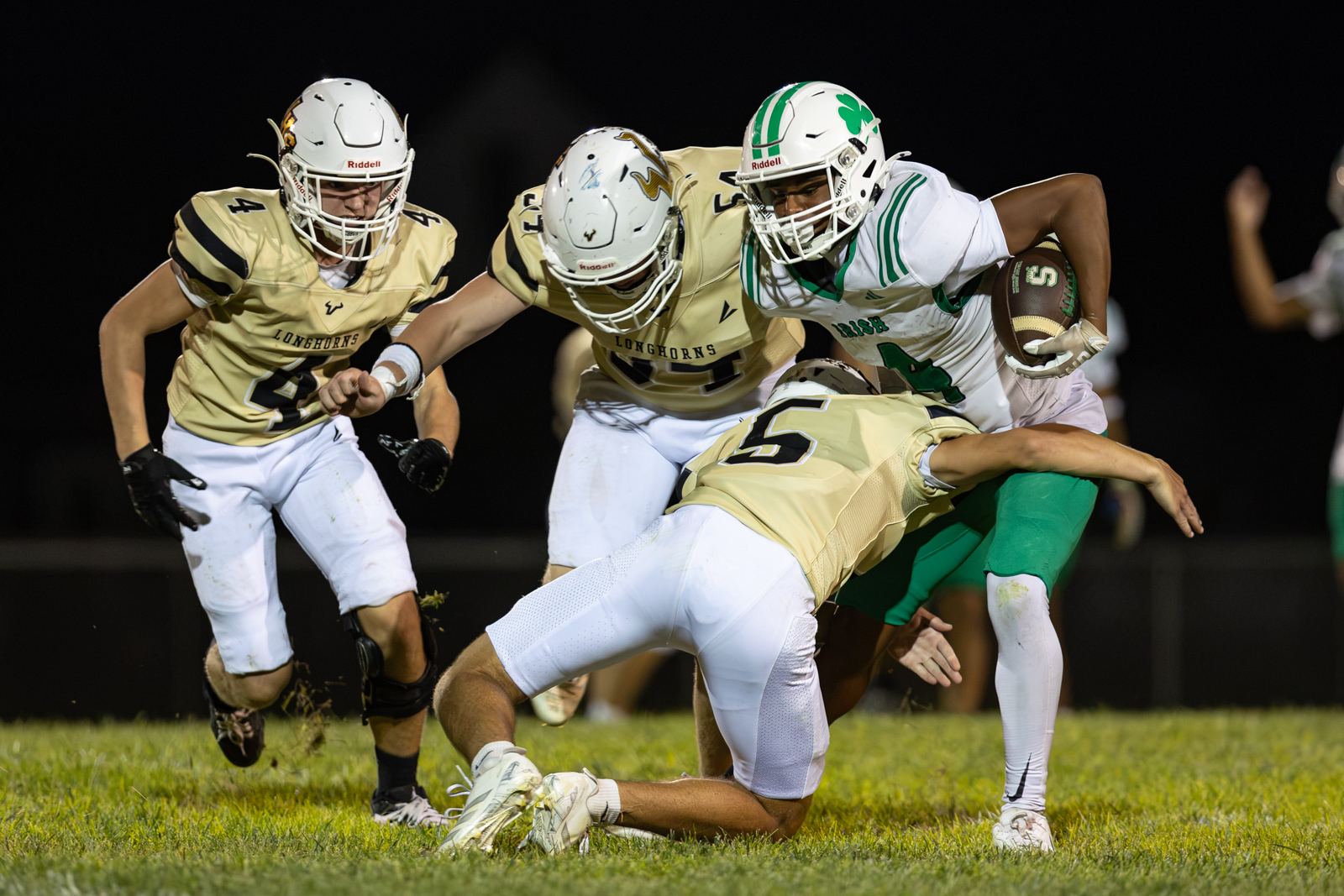 Night football scene: a ball carrier in white and green is tackled by Longhorns players in beige jerseys as they converge on a grassy field under stadium lights.