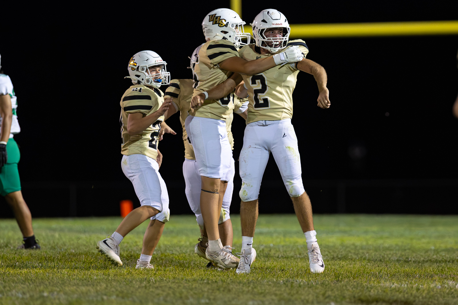 Football players in gold jerseys celebrate a play on a night game, arms raised in triumph on the field.