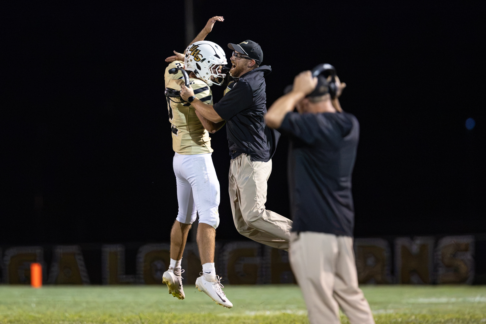 Two American football players in gold jerseys and white pants jump in celebration as a coach embraces them on the sideline during a night game.